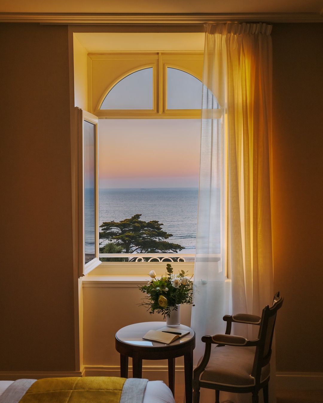Interior shot of a room with a round table and chair in front of an open window showing the ocean and a sunset sky.