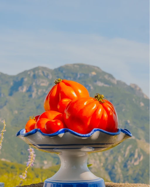 Still life of heirloom tomatoes on a blue and white pedestal dish, set against a mountain landscape and blue sky backdrop.