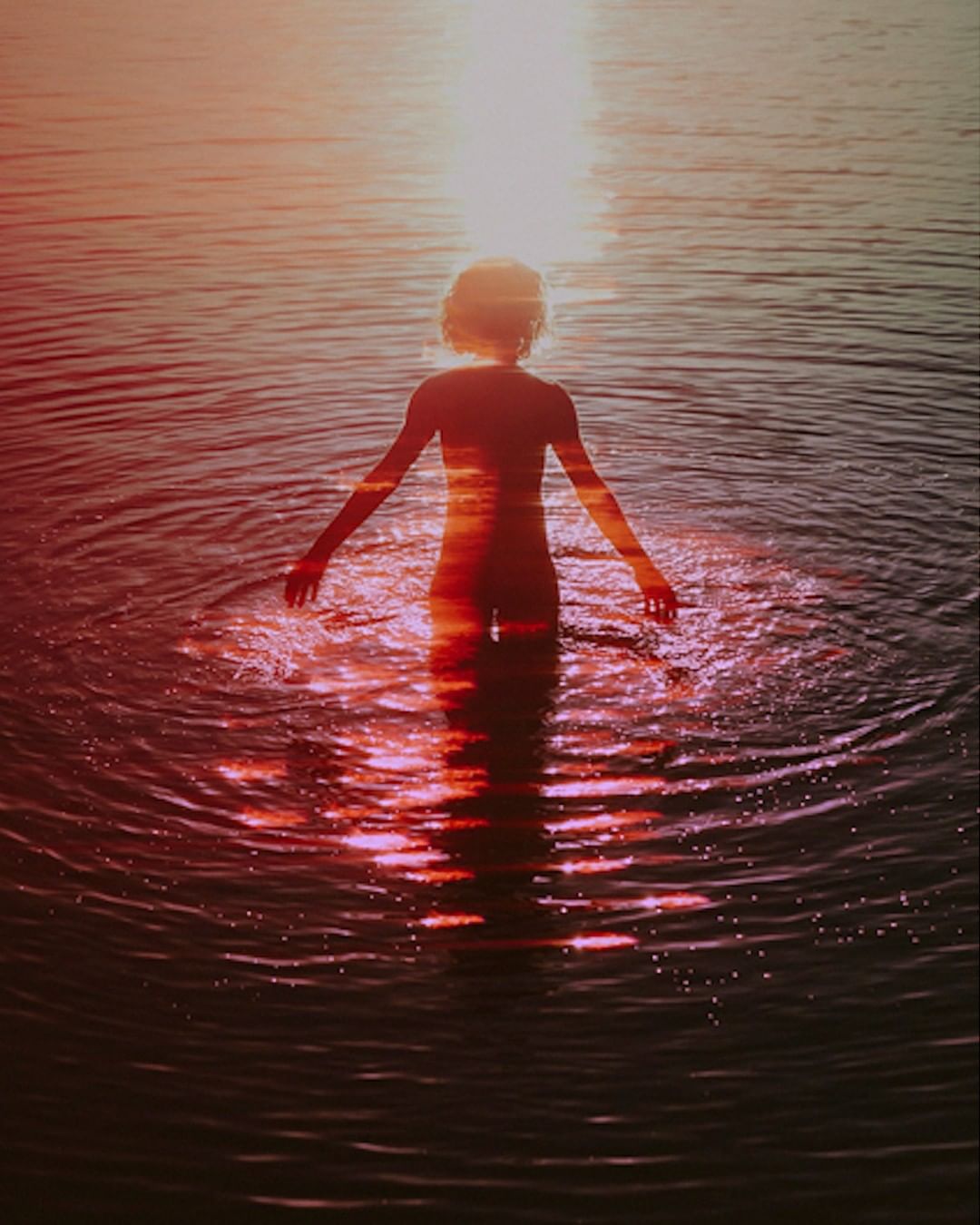 Child stands in water with ripples, backlit by sunset with warm pink and orange tones creating a dreamy, surreal atmosphere.