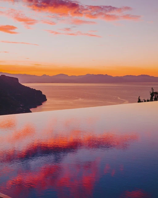 Scenic view of sea and mountains with reflections in infinity pool at sunset, featuring orange and purple sky and calm water.