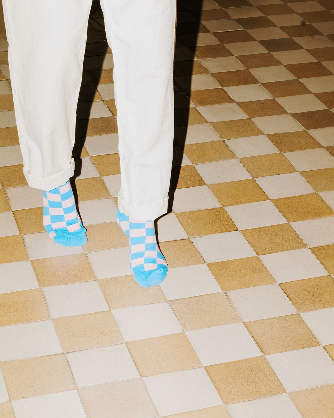 Person in white pants and blue checkered socks standing on a tan and white checkered tile floor in a brightly lit indoor space.