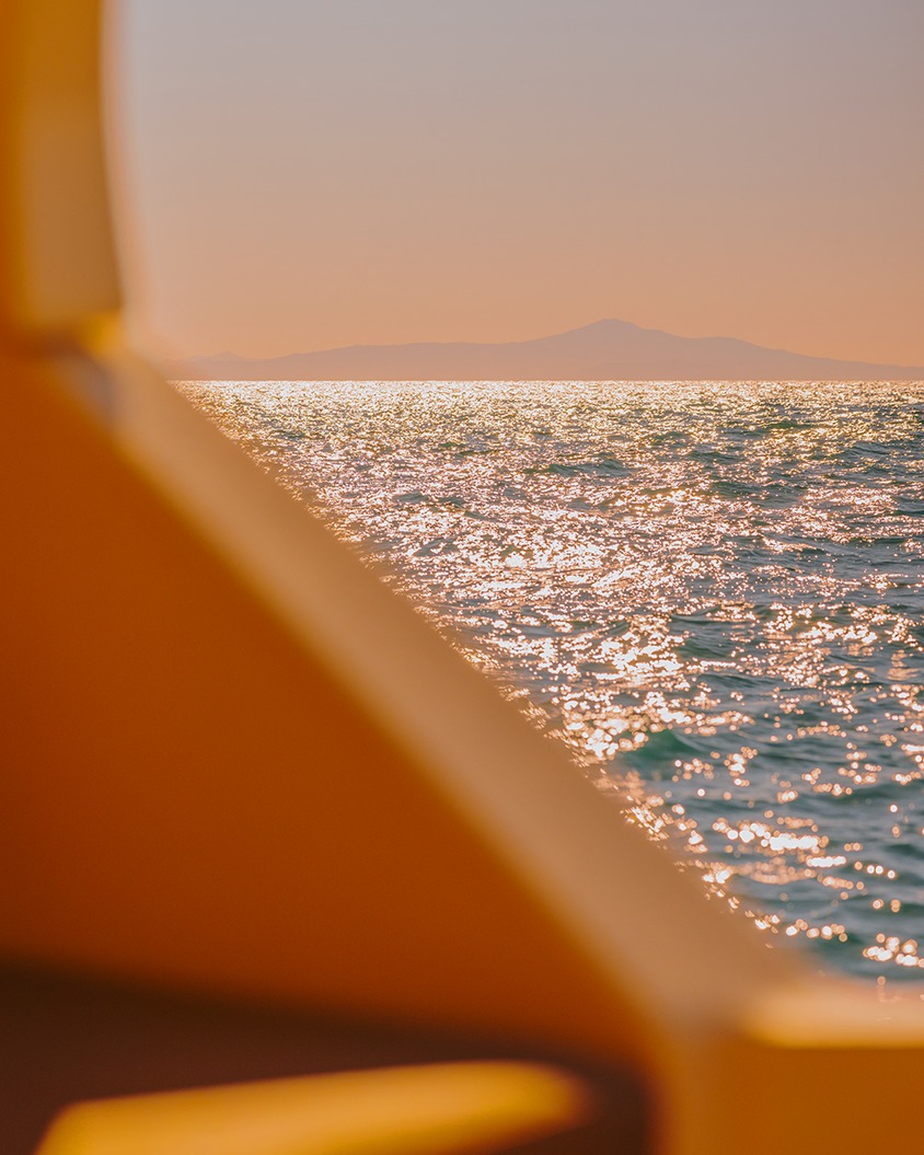 Sparkling water surface reflects sunlight, seen through a boat railing with distant mountains and peach sky in background.