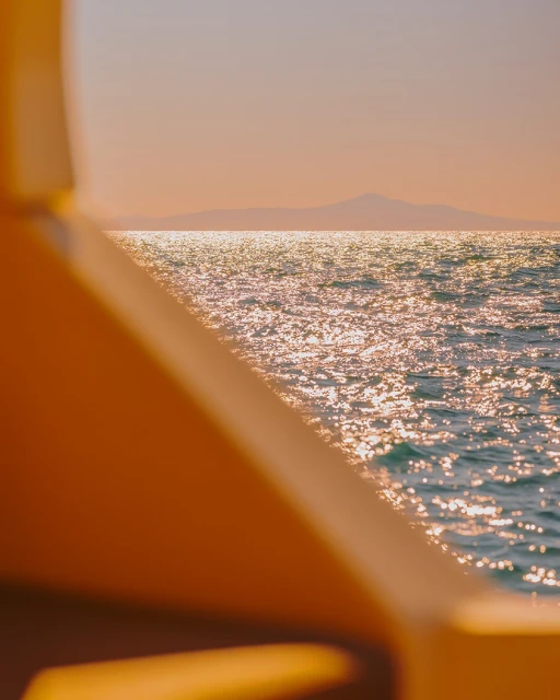 Sparkling water surface reflects sunlight, seen through a boat railing with distant mountains and peach sky in background.