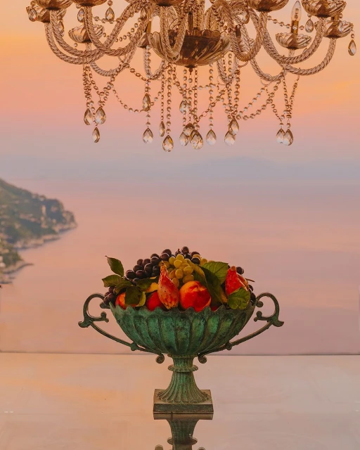 Still life with a fruit bowl beneath an ornate chandelier, set against a scenic coastal background at sunset.