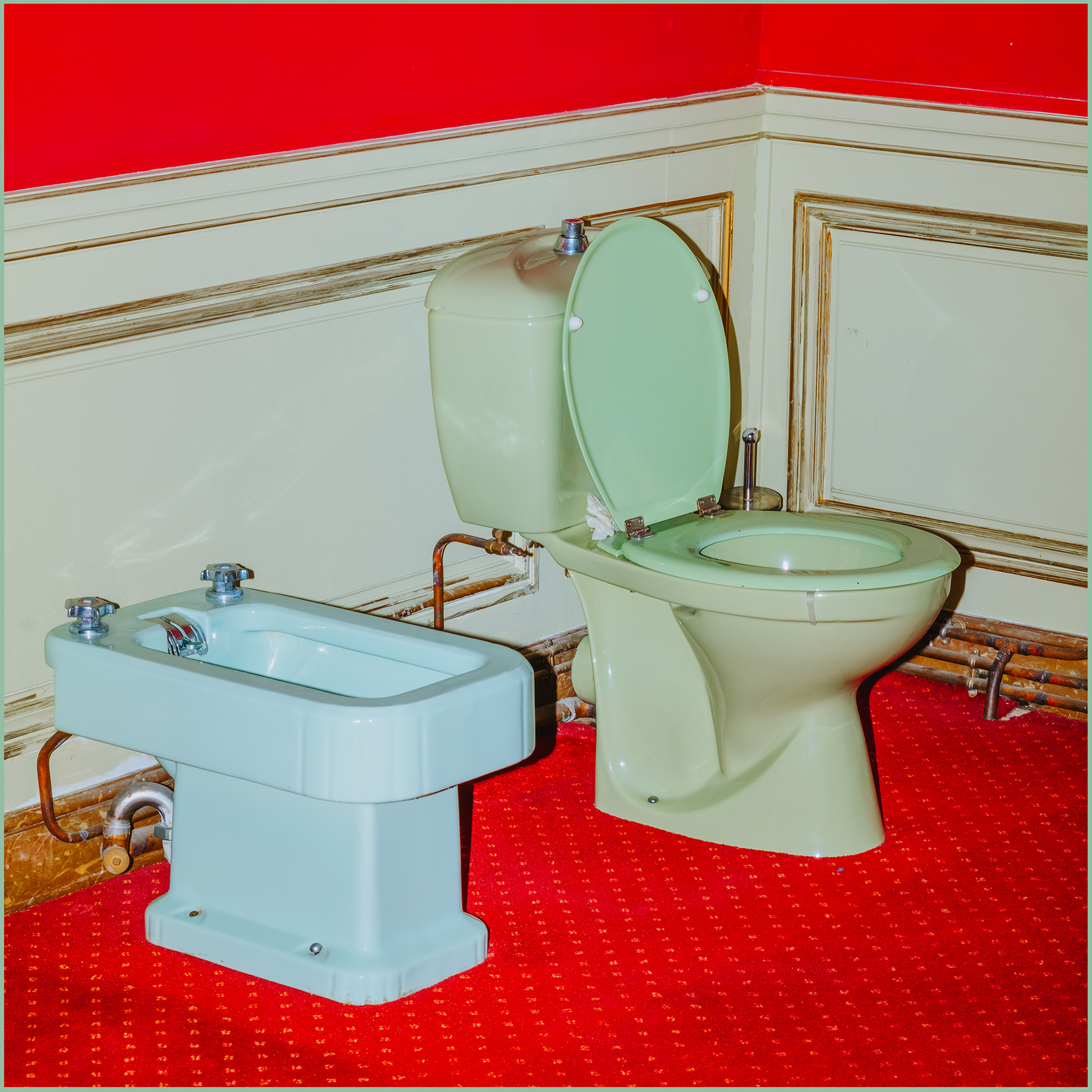 Retro-style bathroom interior showcasing a green toilet and blue bidet against a red floor and red-and-cream wall.