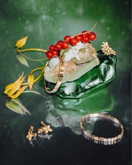 Fine jewelry including a gold bracelet, ring, and earrings displayed on stone with berries and flowers against a green background.