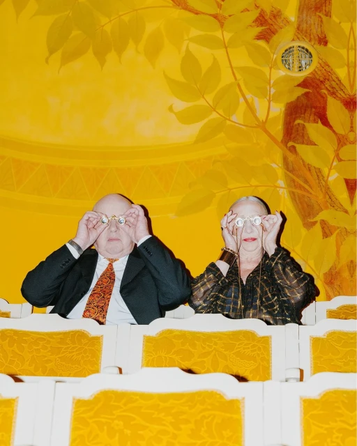 Elderly couple in theatre box seats look through opera glasses. Ornate gold details decorate the balcony seats and wall mural.