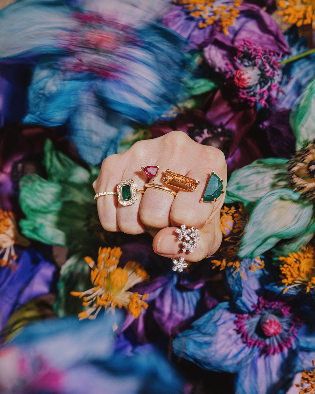 Hand wearing multiple rings, including emerald and ruby rings, set against a background of blue, purple, and orange flowers.