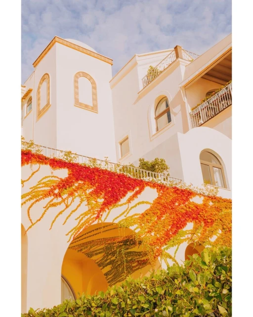 Exterior of a white stucco building with archways, windows, and a balcony, adorned with red and orange vine foliage under a blue sky.
