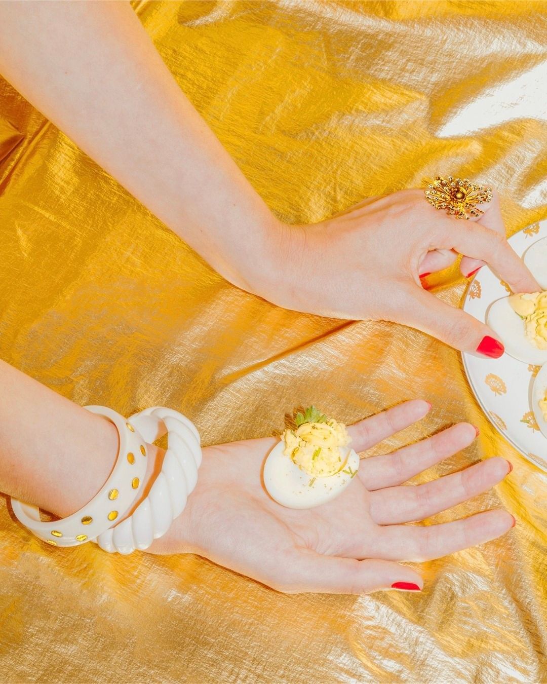Overhead shot featuring a woman's hands, adorned with a large ring and bracelets, holding a deviled egg against a shimmering gold background.