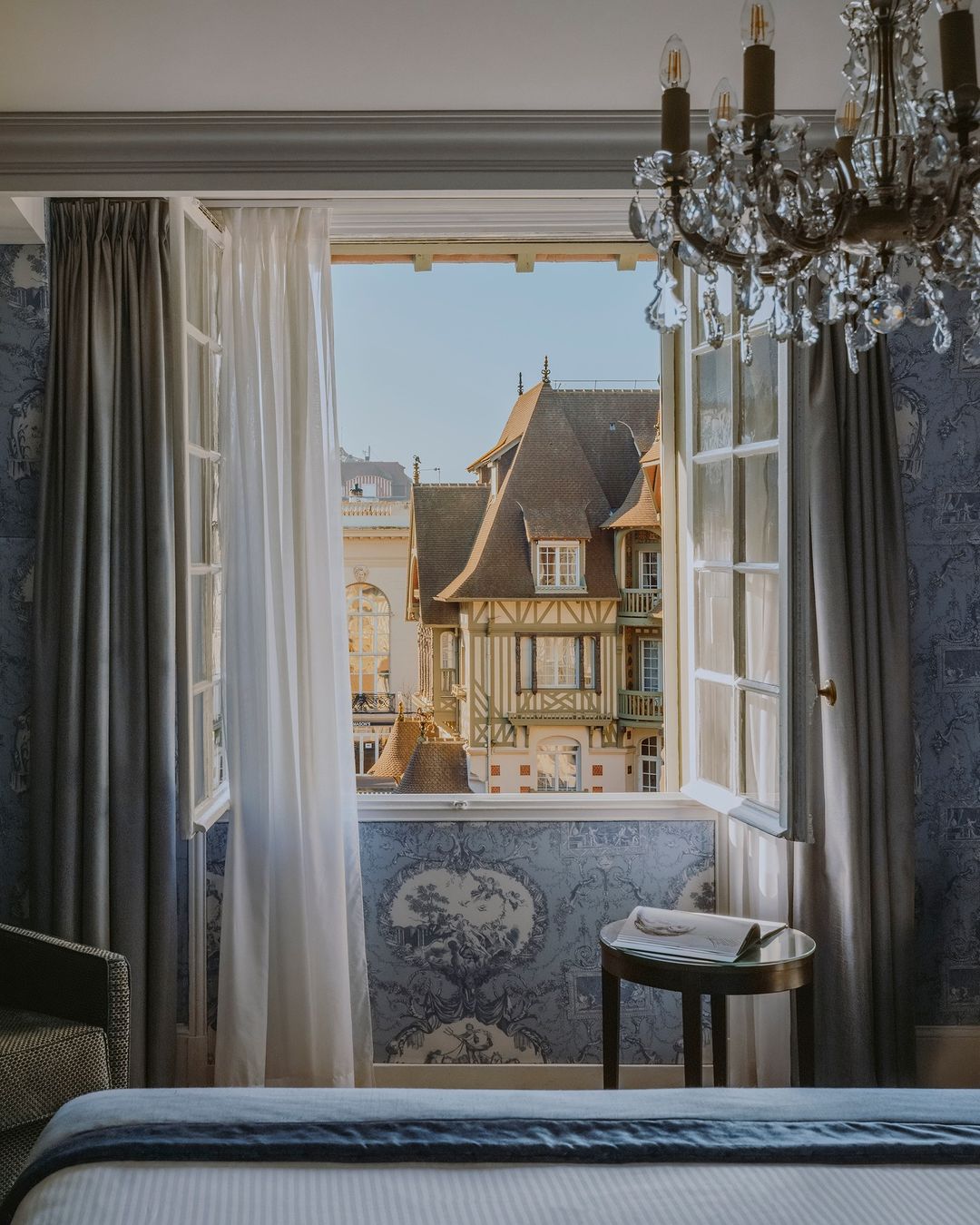 Bedroom interior view through open window of European-style timbered building, curtains, chandelier, and patterned wallpaper.