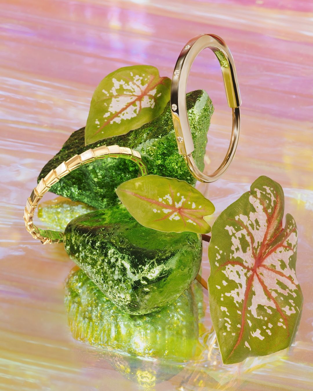 Gold bracelets styled on green rocks and leaves against a pink iridescent background, jewelry product shot for fashion and beauty.