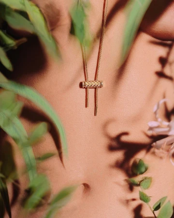 Elegant close-up of a woman's torso wearing a gold braided pendant necklace amidst blurred green foliage for a natural aesthetic.
