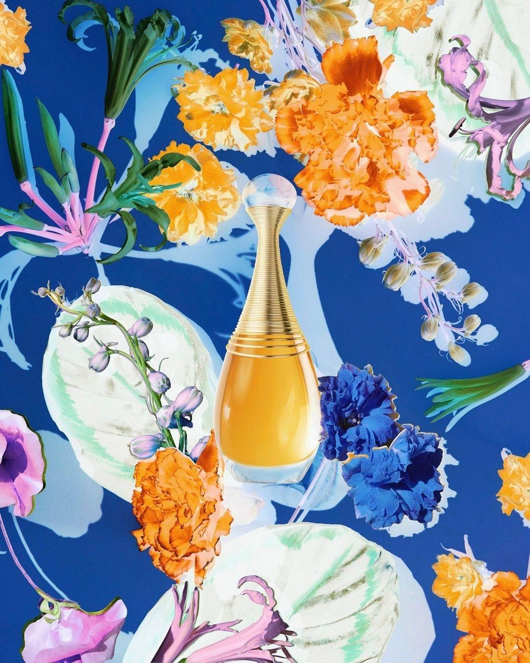 Gold perfume bottle on a blue background with orange and blue flowers and green leaves, creating an artistic beauty product shot.