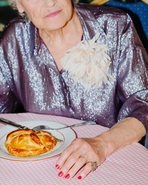 Elderly woman in a shiny purple shirt sits at a table with a pastry on a plate. She has a white feather brooch and red nails.