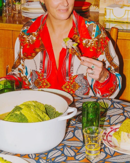 Adult woman in patterned shirt at a table eating stuffed cabbage from a white bowl, with green drinking glasses and patterned tablecloth.