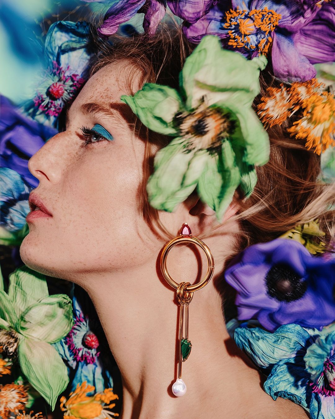 Profile view of a woman surrounded by flowers with artistic makeup, gold earring, and fair skin, showcasing beauty and floral design.