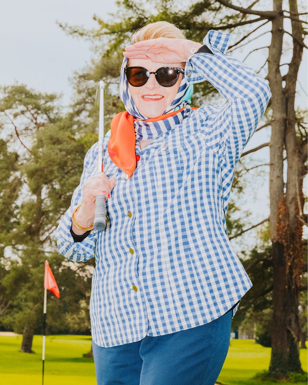 Older woman in sunglasses and headscarf on golf course shields eyes from sun, holding golf club, wearing blue gingham shirt.