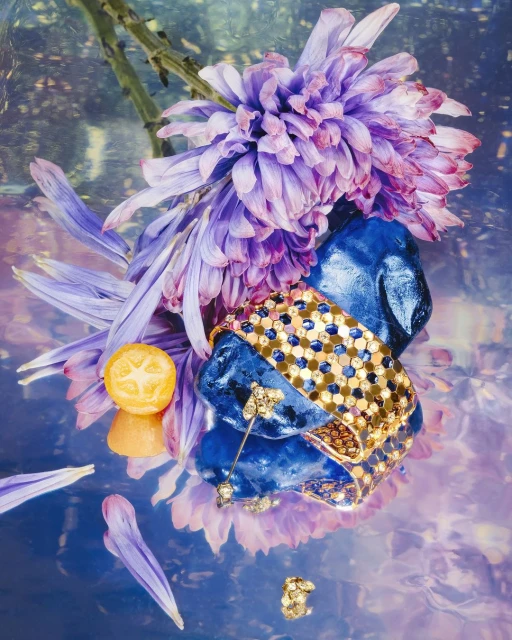 Still life arrangement featuring a gold bracelet draped over blue stones, alongside purple flowers and a sliced cumquat, on a reflective surface.