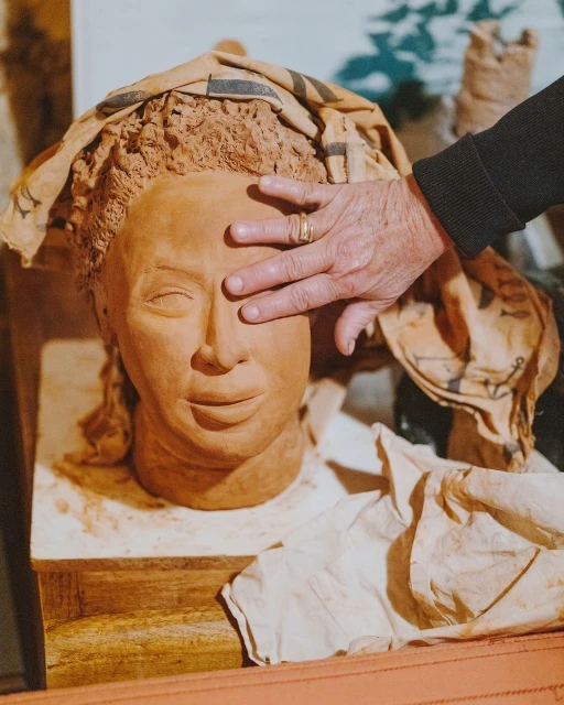 Sculpture of a head with a cloth covering the hair and a person's hand covering one eye. The sculpture rests on a wooden surface.
