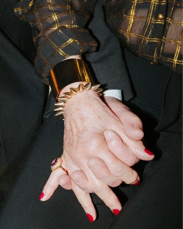 Close-up of an elderly couple holding hands, showing a gold spiked cuff bracelet, gold ring with a purple gem, and red nail polish.