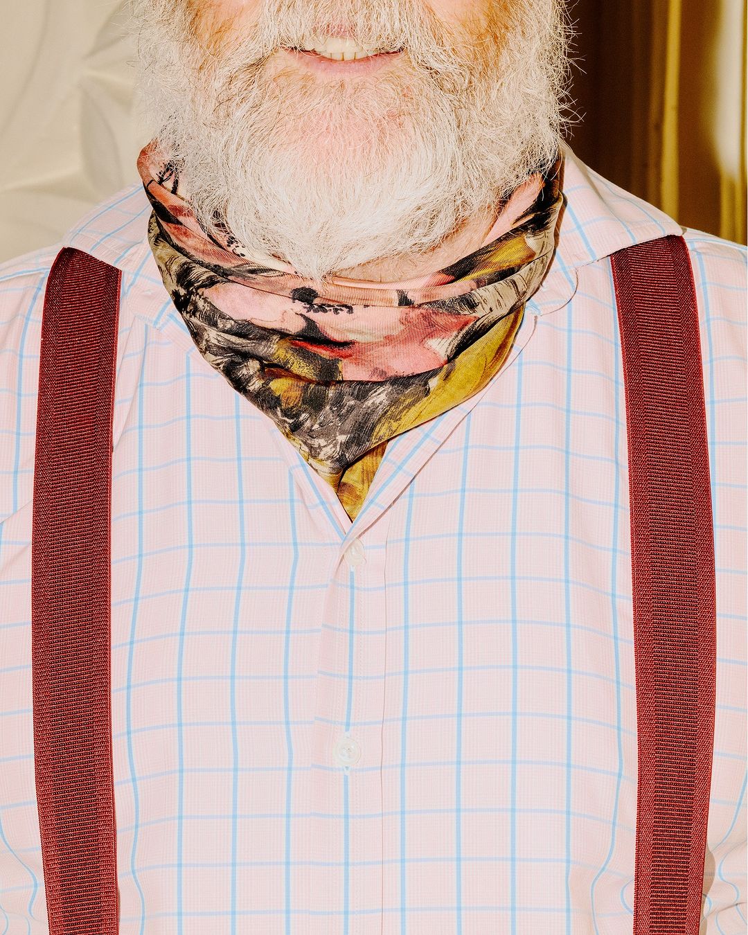 Close-up of adult man with white beard, wearing a pink checkered shirt with suspenders and a patterned scarf, smiling softly.