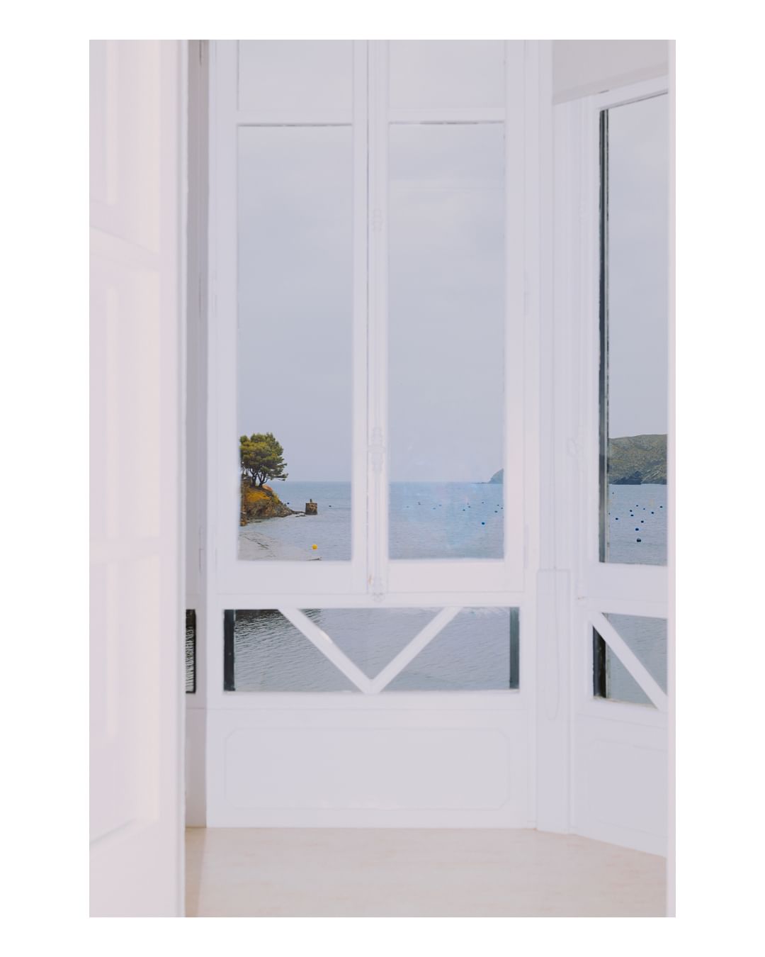 View from a bright, white, interior through an open multi-pane window to a serene coastal landscape with a small island and blue sea.