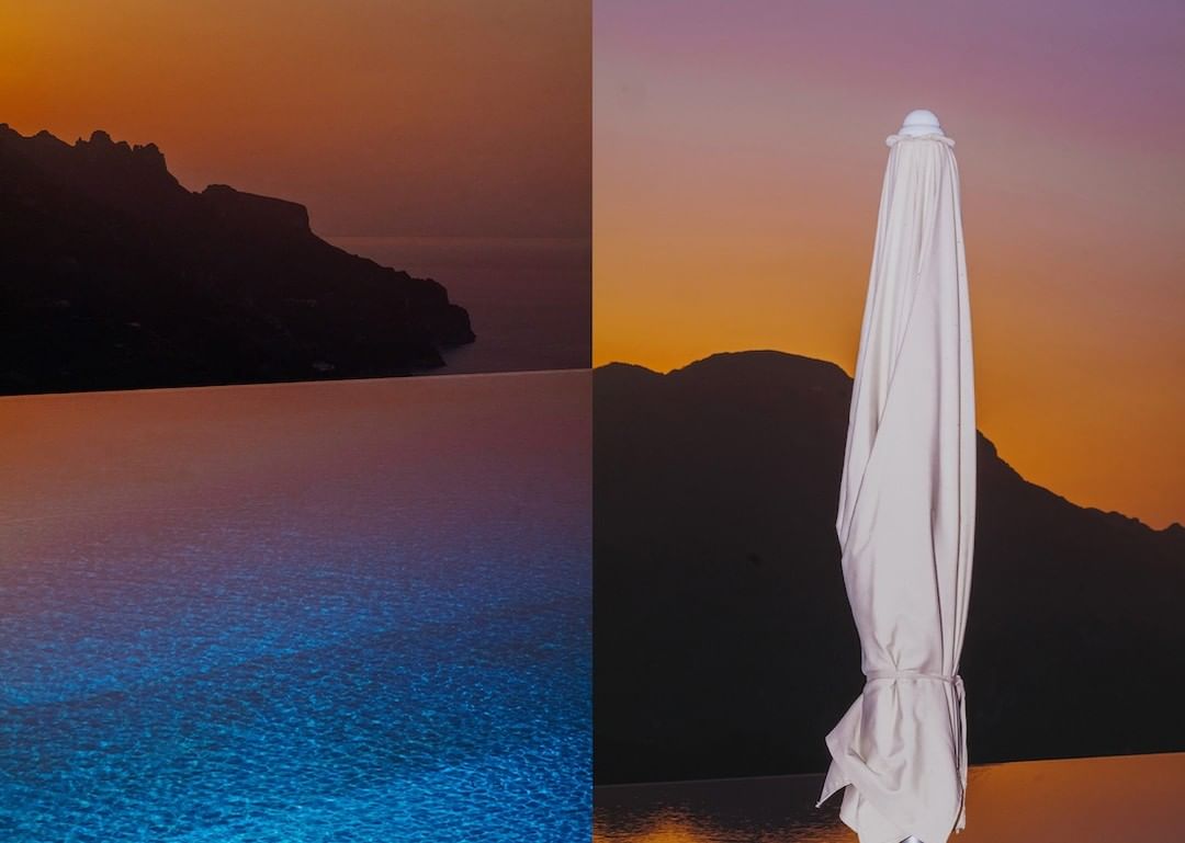 Landscape featuring an infinity pool blending into the ocean at sunset, with mountains in the background and a closed white umbrella.