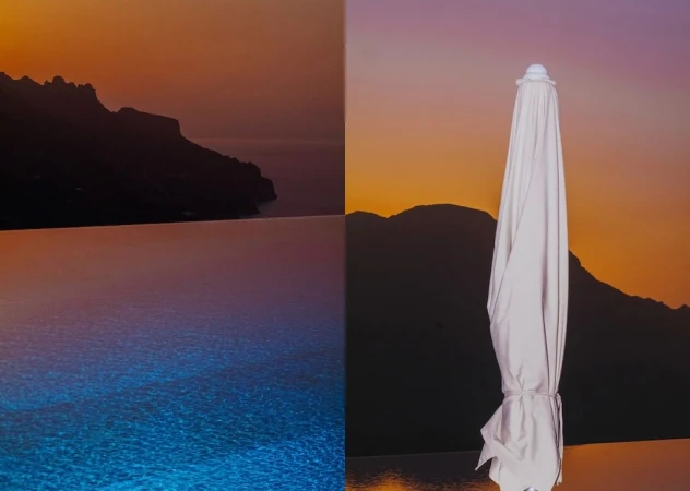 Landscape featuring an infinity pool blending into the ocean at sunset, with mountains in the background and a closed white umbrella.