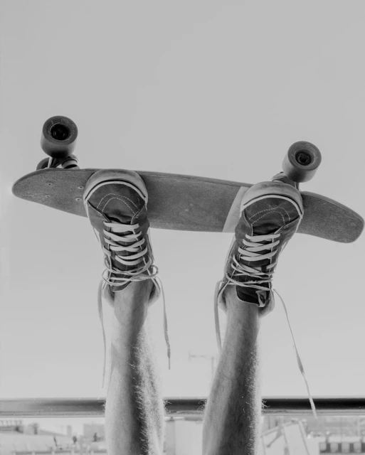 Black and white shot of an adult's legs lifted with feet on a skateboard, against a background with buildings and bright sky.