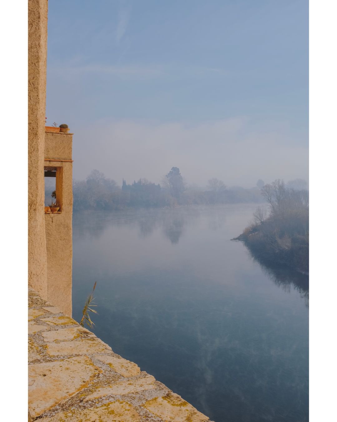 Scenic view of a river under a misty sky, framed by an old building on the left side, evoking a sense of serenity and timelessness.