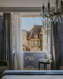 An open window in a luxury hotel room reveals a historic timbered building exterior and blue sky in Paris.
