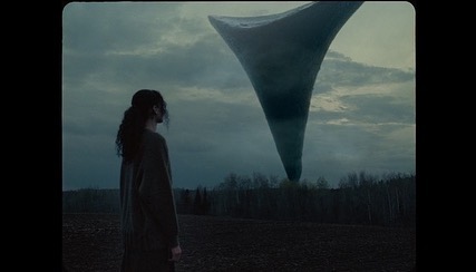 A woman stands in a field at dusk, gazing up at a large, inverted cone shape in the sky, with bare trees visible in the distance.