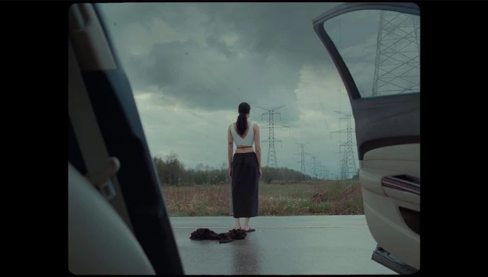 Woman standing with her back to the camera on an open road viewed through a car door opening under an overcast sky.
