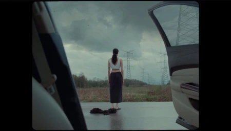 Woman standing with her back to the camera on an open road viewed through a car door opening under an overcast sky.