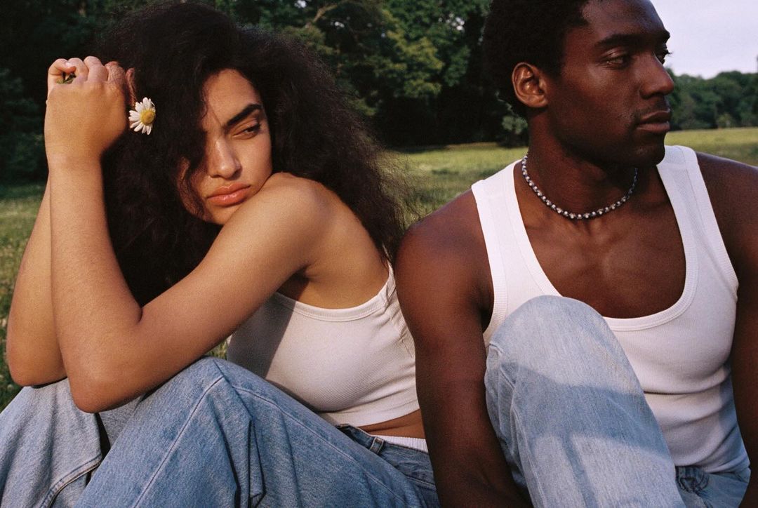 A stylish biracial couple poses in a grassy field, wearing casual tank tops and jeans in warm sunlight, showcasing natural beauty and style.