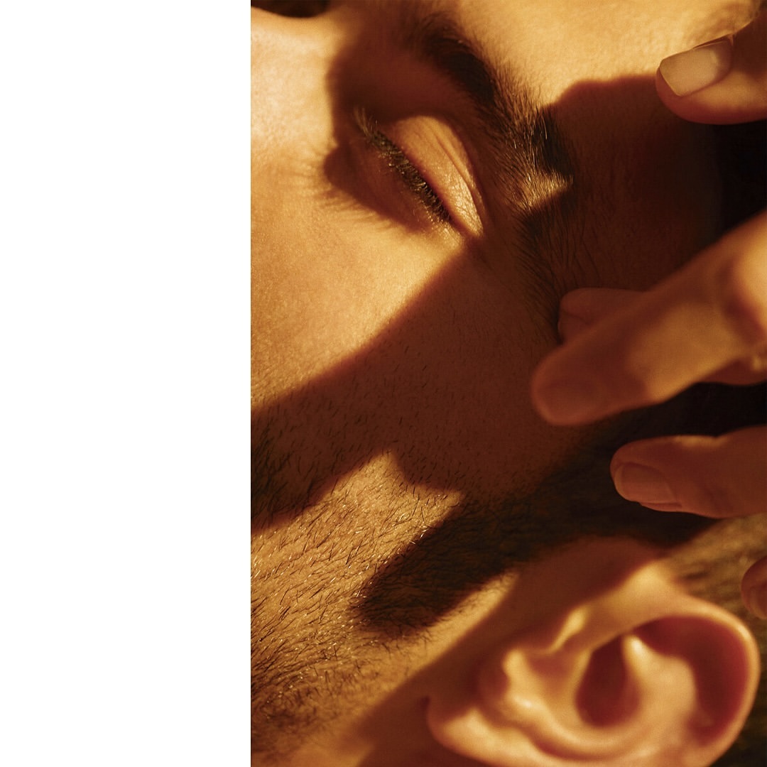 Close-up portrait of an adult male's face, featuring tanned skin and the partial view of hands casting a shadow over his face.