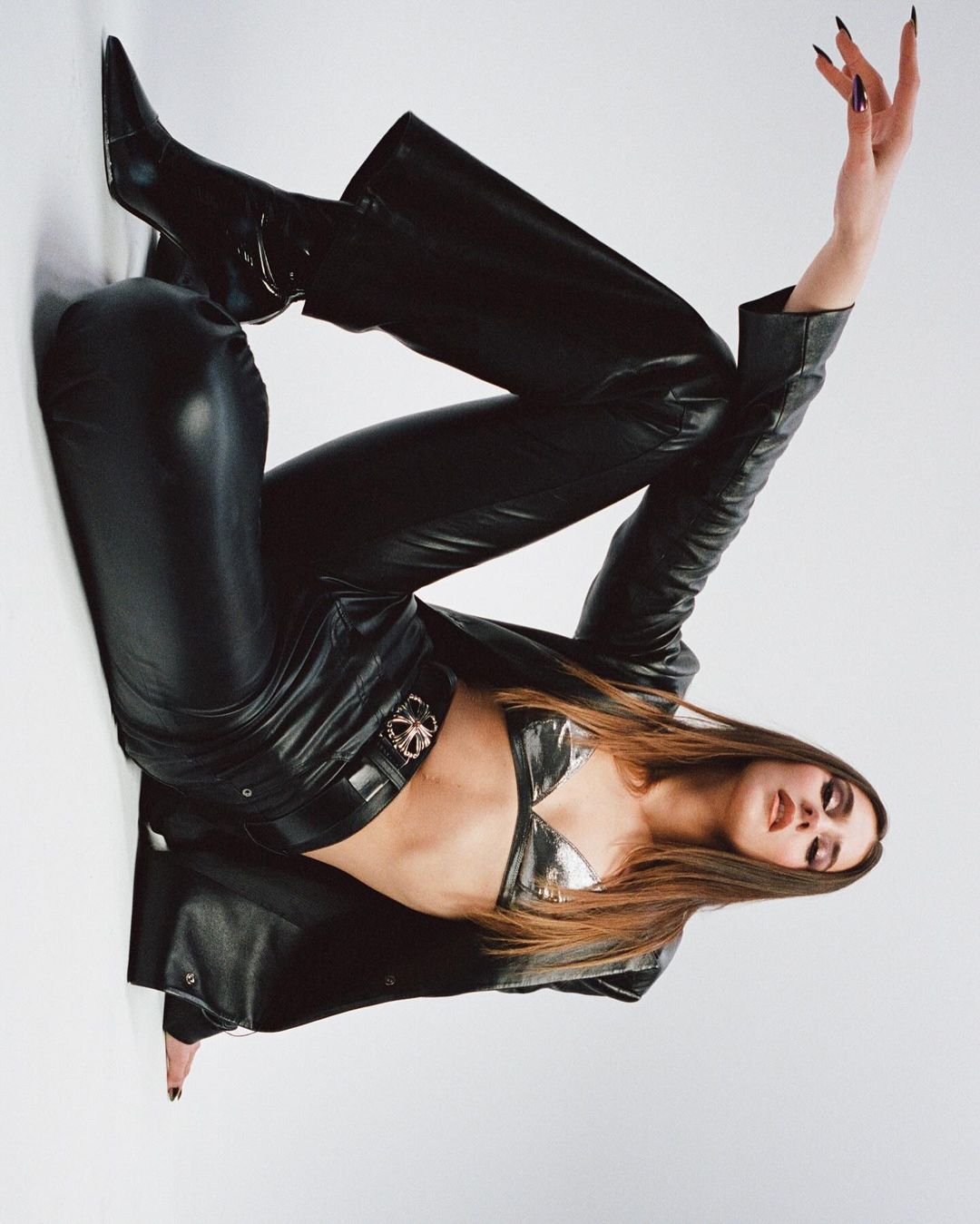 An adult female with long brown hair poses in a photo studio wearing a metallic bra top, black leather jacket, pants and boots on white background.