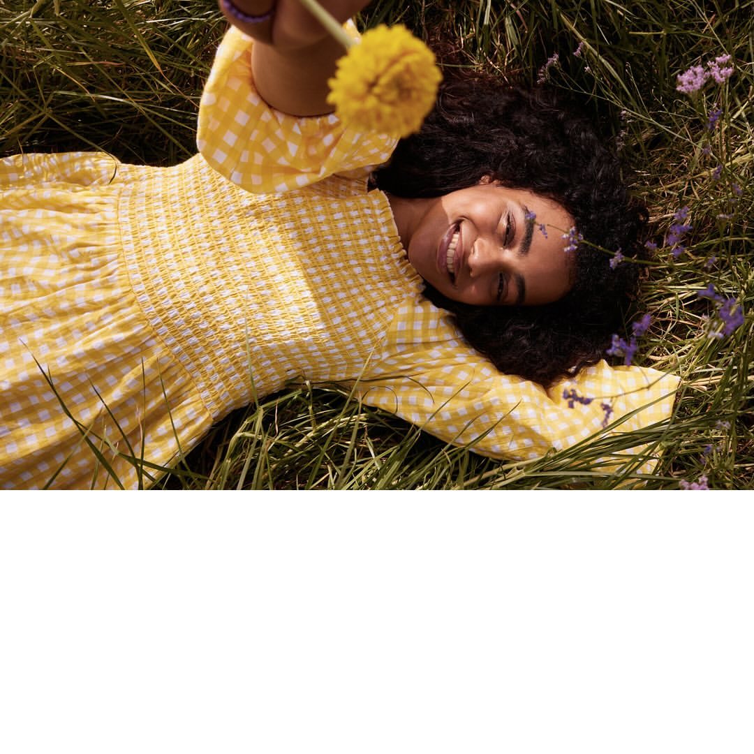 Woman in yellow gingham dress lies on grass, holding dandelion flower, smiling in outdoor setting with a cheerful, summery vibe.