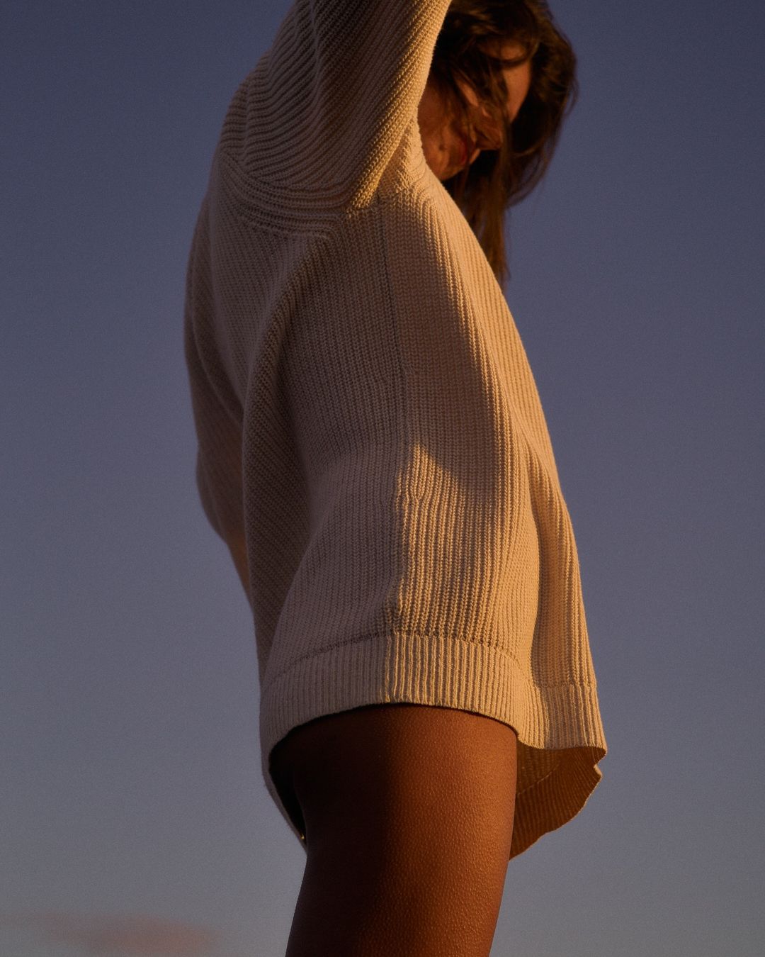 Woman in oversized knit sweater with arms raised against a dusk sky; low-angle view highlighting clothing texture and the evening sky.
