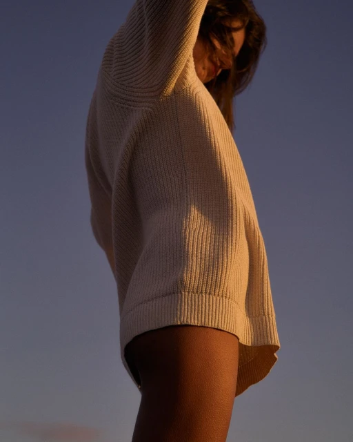 Woman in oversized knit sweater with arms raised against a dusk sky; low-angle view highlighting clothing texture and the evening sky.