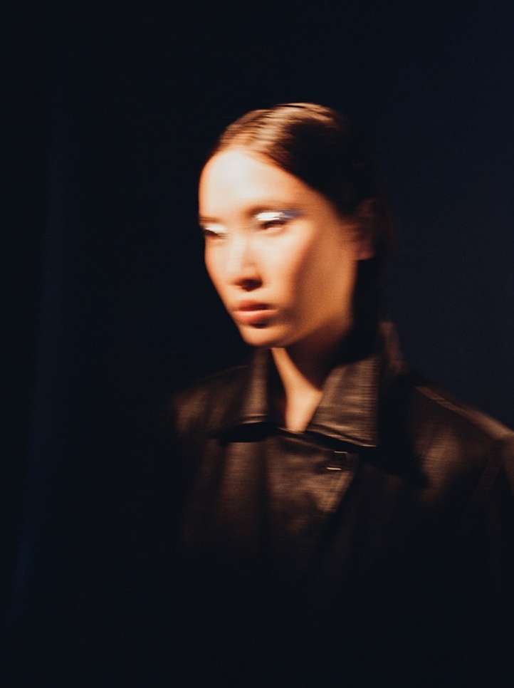 Portrait of a beautiful Asian woman with dramatic white makeup and a leather jacket against a dark background in a studio setting.
