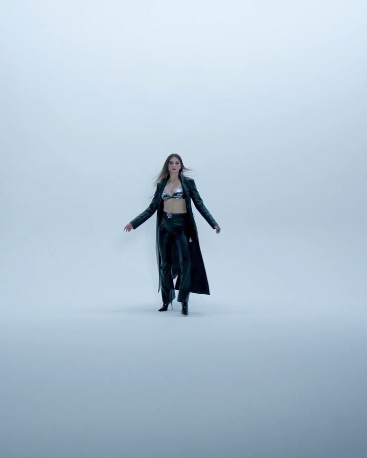 Fashion shot of an adult female model in a metallic bra top, black leather pants, heels, and a long coat, posing in a studio setting.