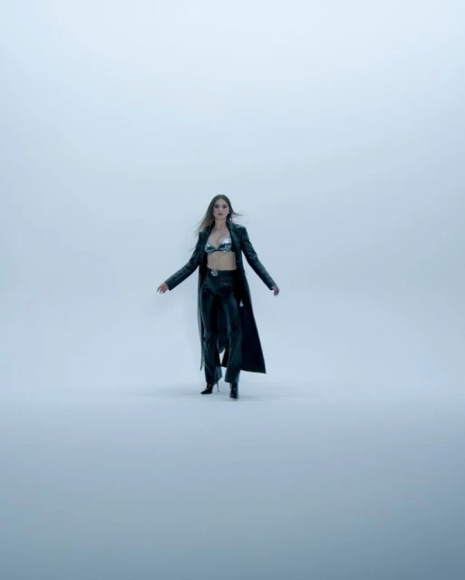 Fashion shot of an adult female model in a metallic bra top, black leather pants, heels, and a long coat, posing in a studio setting.
