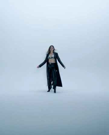 Fashion shot of an adult female model in a metallic bra top, black leather pants, heels, and a long coat, posing in a studio setting.