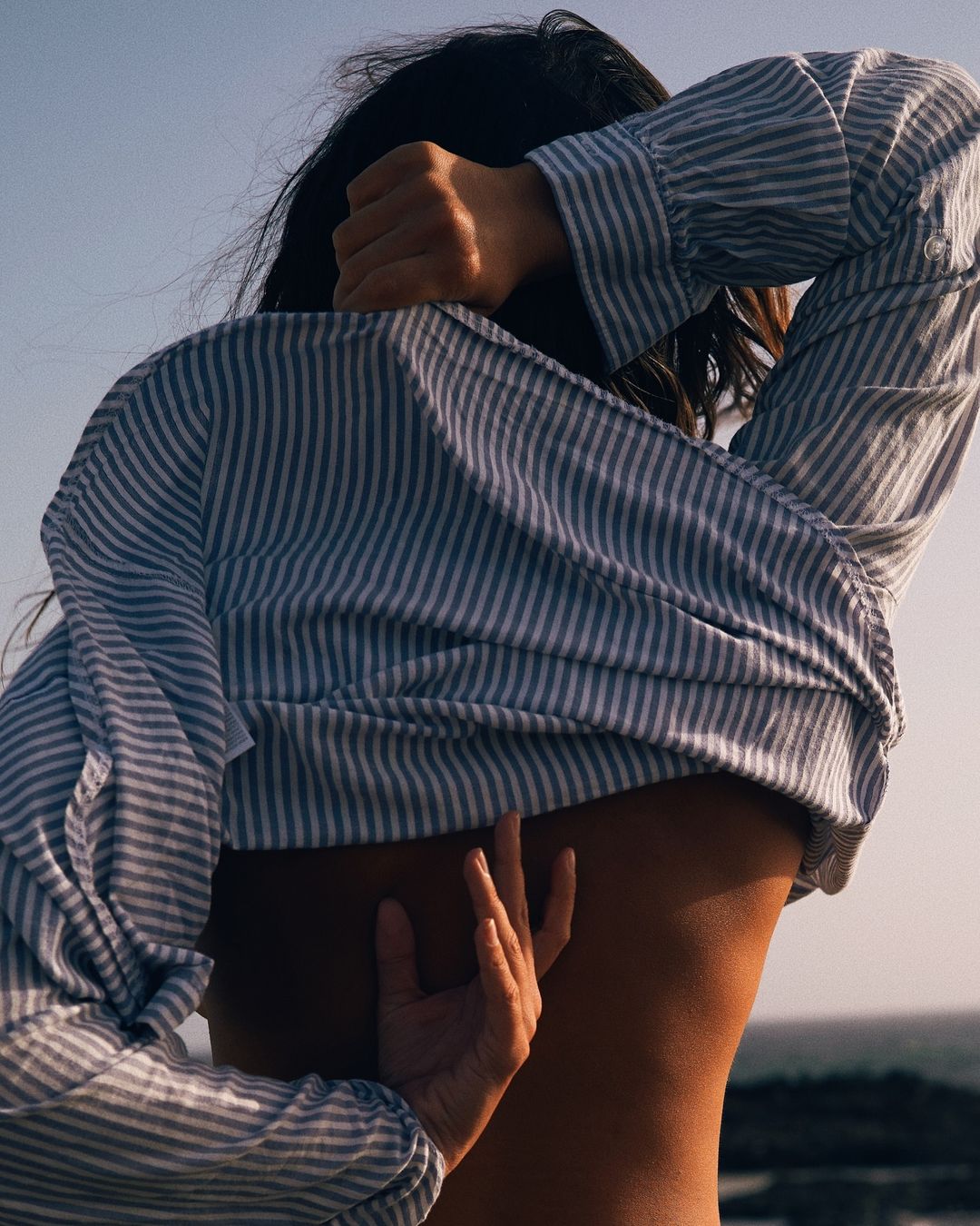 Back view of a slender adult woman in a striped shirt with her hand gently touching her back outdoors during daytime.
