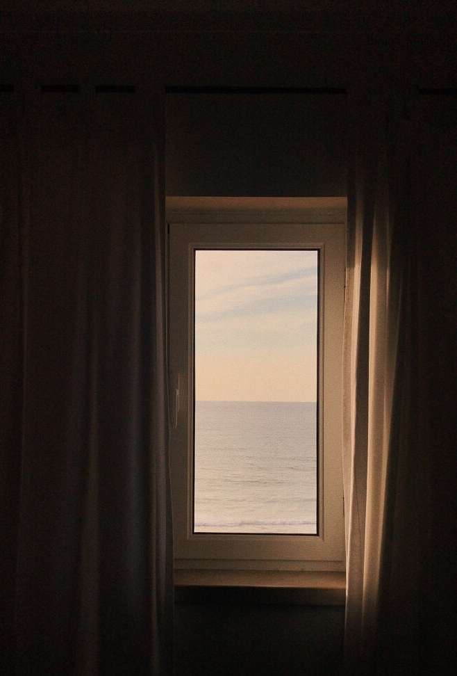 View of the ocean through a white-framed window, flanked by heavy beige curtains, creating a cozy, inviting atmosphere.
