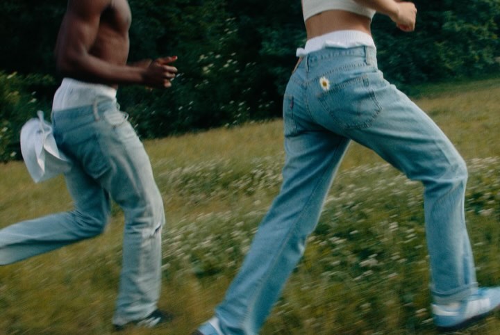 Two people run through a grassy field with scattered white flowers, wearing light blue jeans and athletic shoes in a daytime outdoor scene.