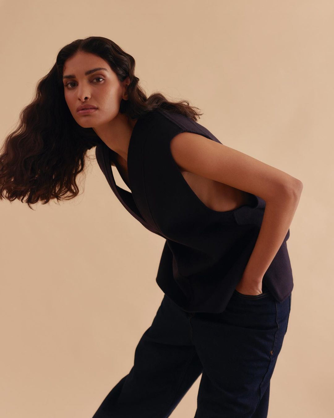 Woman in a studio shot wearing a black top and jeans, leaning forward with a serious expression, against a cream-colored backdrop.