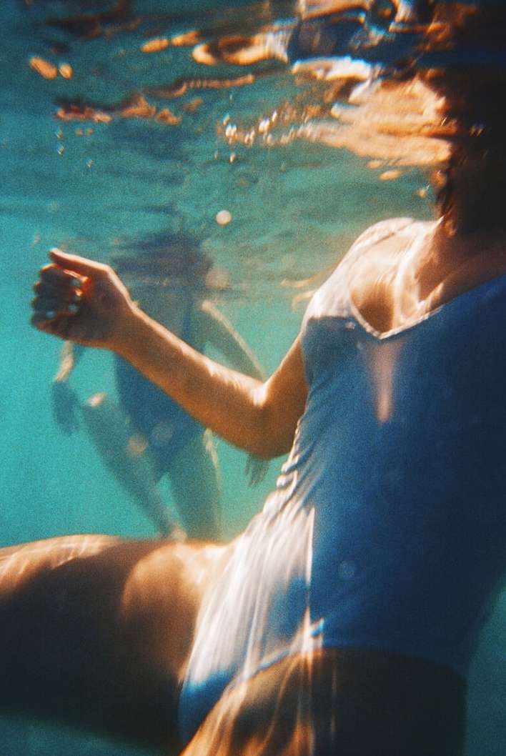 Two people wearing blue swimsuits are swimming underwater in clear, light-blue water, creating a serene and dreamlike aquatic scene.